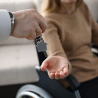 Zbliżenie na biznesmena wręczającego klucze od nowego domu niepełnosprawnej kobiecie. Kobieta siedząca na wózku inwalidzkim. Dzień przeprowadzki do mieszkania. Niepełnosprawni i koncepcja nieruchomości